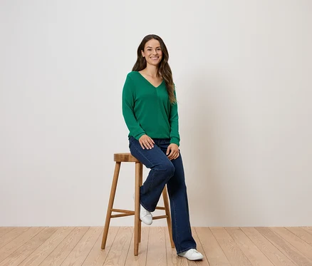 Une femme est assise sur un tabouret en bois, portant un pull vert et un jean.