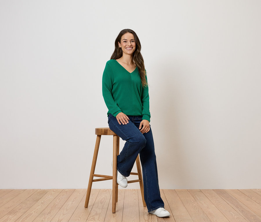Une femme est assise sur un tabouret en bois, portant un pull vert et un jean.