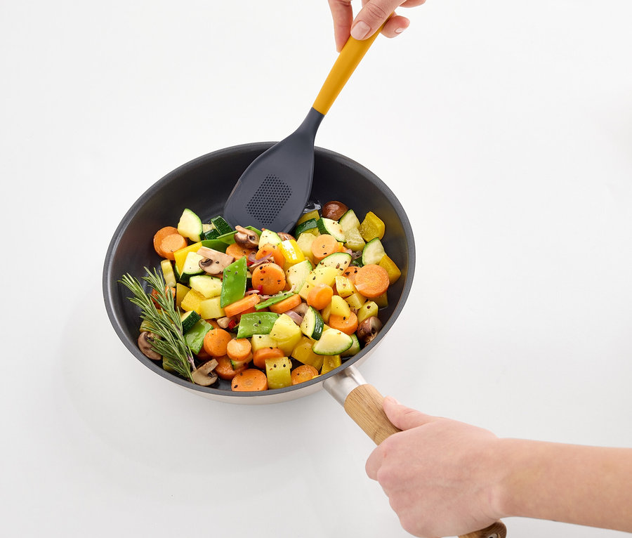 Des mains mélangent des légumes dans une poêle avec une spatule ajourée jaune.
