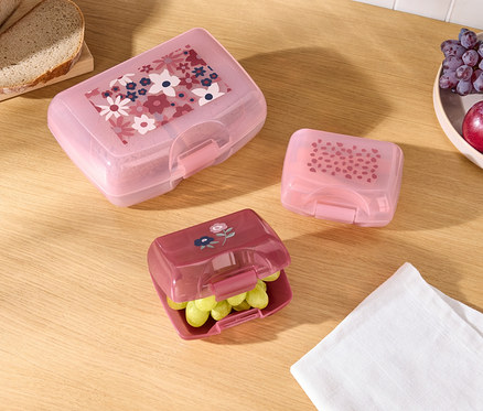 Trois boîtes à goûter colorées avec différents motifs, du pain et des fruits sur une table en bois.