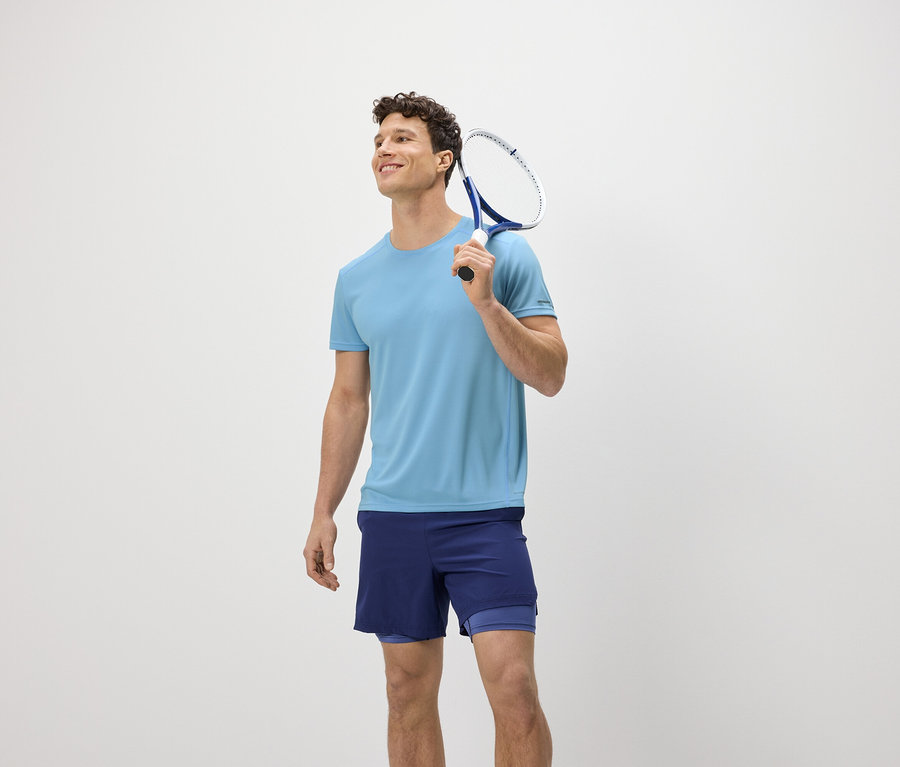 Un homme avec une raquette de tennis sur l'épaule porte un t-shirt fonctionnel bleu clair.