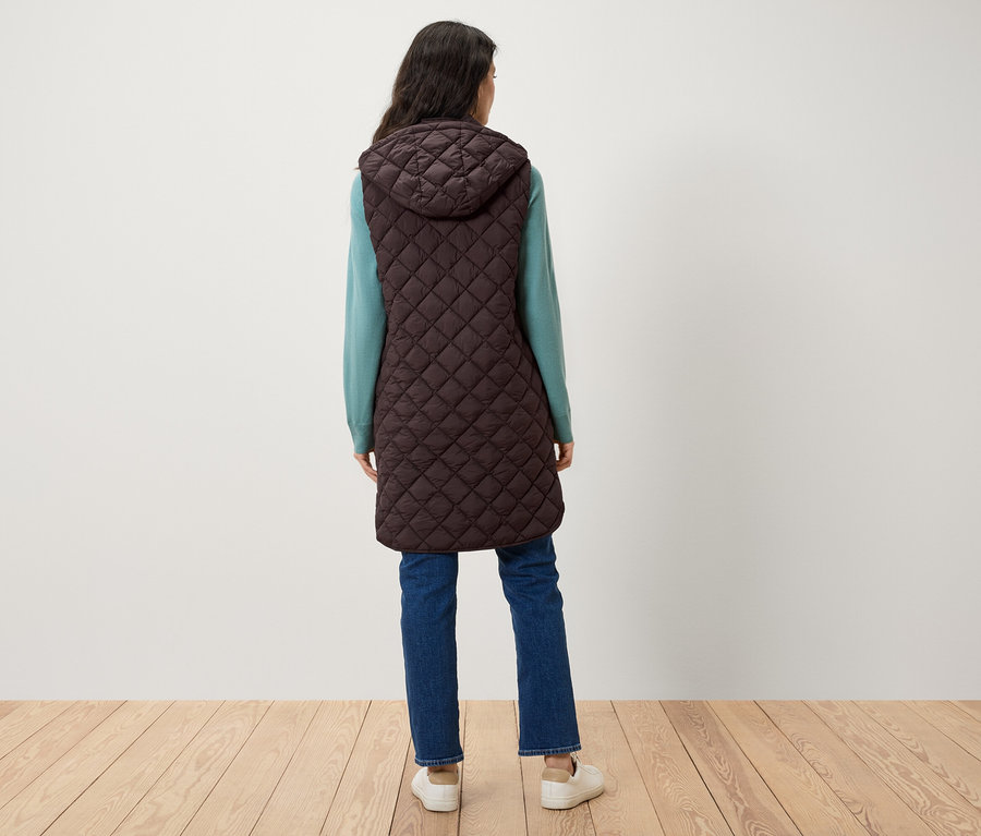Une femme se tient dos à l'objectif, portant un gilet matelassé marron à capuche, un pull bleu et un jean.