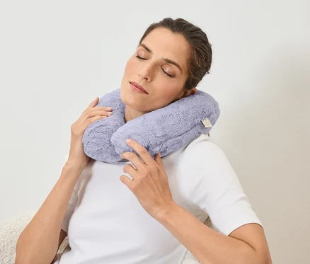 Une femme les yeux fermés tient un coussin de voyage en peluche violet clair autour de son cou avec ses mains.