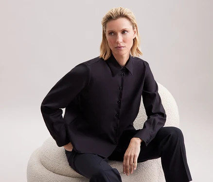 Une femme blonde est assise sur un fauteuil beige et porte une chemise noire et un pantalon.