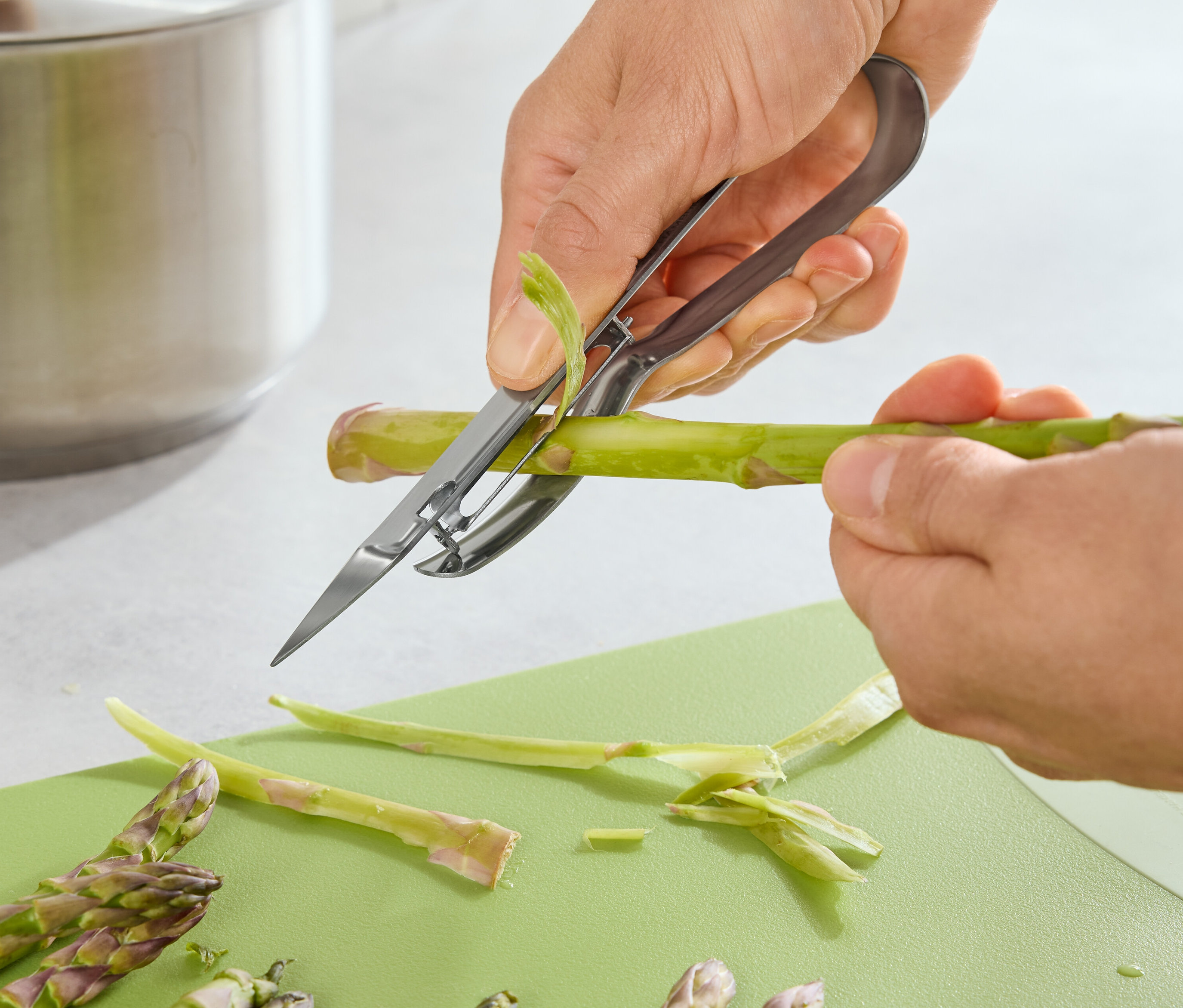 Une personne pèle des asperges avec un éplucheur à asperges sur une planche à découper verte.
