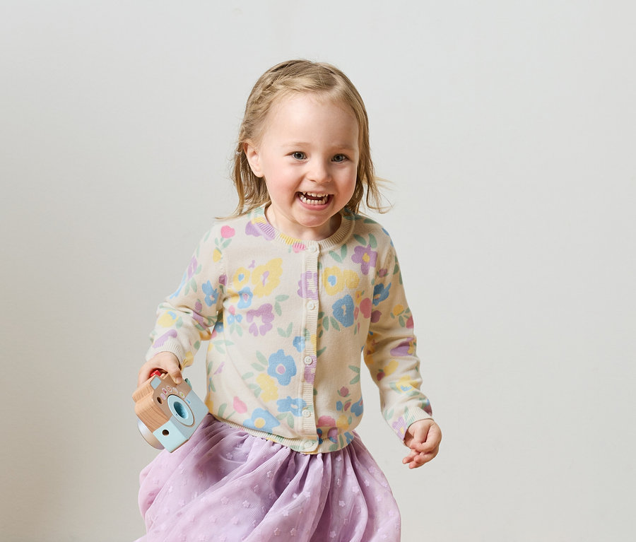 Fillette blonde souriante avec des tresses, un cardigan à fleurs et une jupe violette tenant un appareil photo jouet.