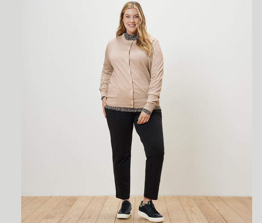 Une femme porte une blouse imprimée à col montant, un cardigan en maille fine et un pantalon de jogging noir.