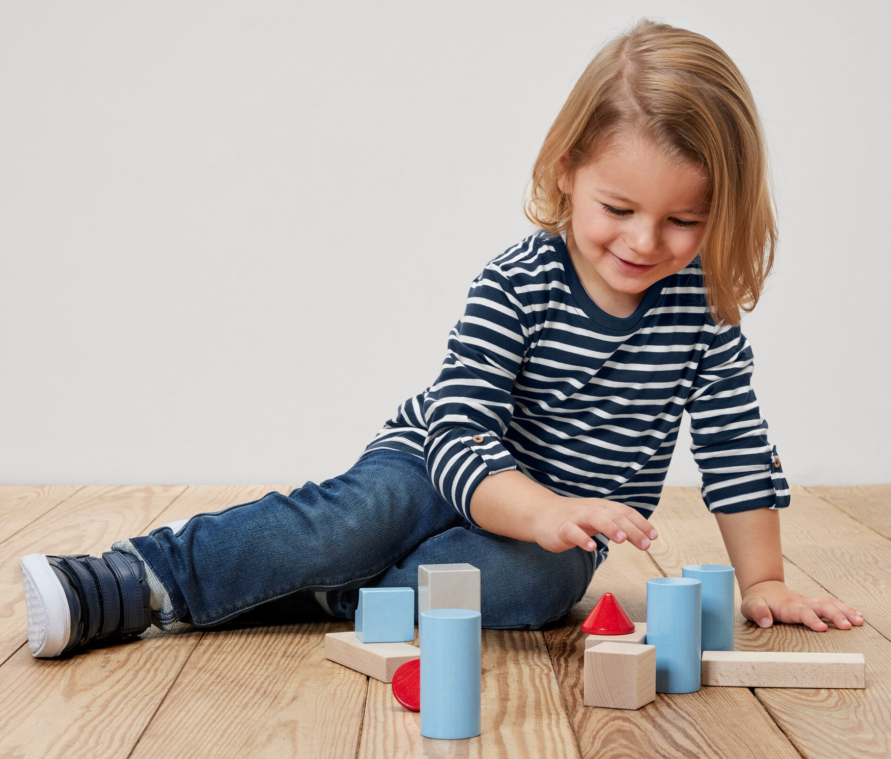 Un enfant est assis par terre et joue avec des blocs de bois colorés. Il porte 2	-shirts manches longues pour enfant.