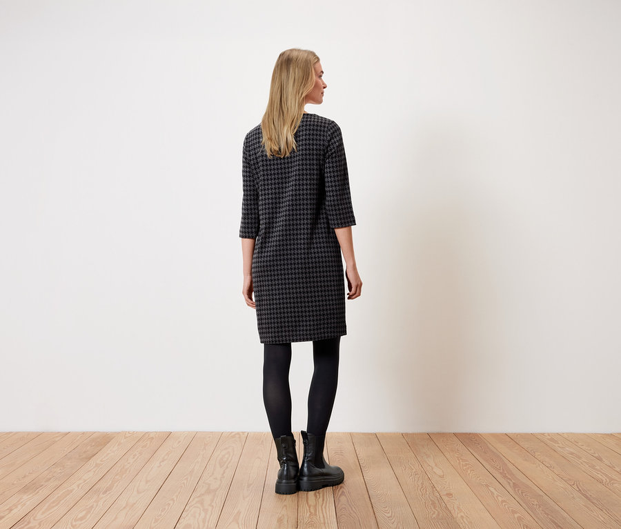 Une femme se tient dos à la caméra et porte une robe à motif pied-de-poule, des leggings noirs et des bottes noires.