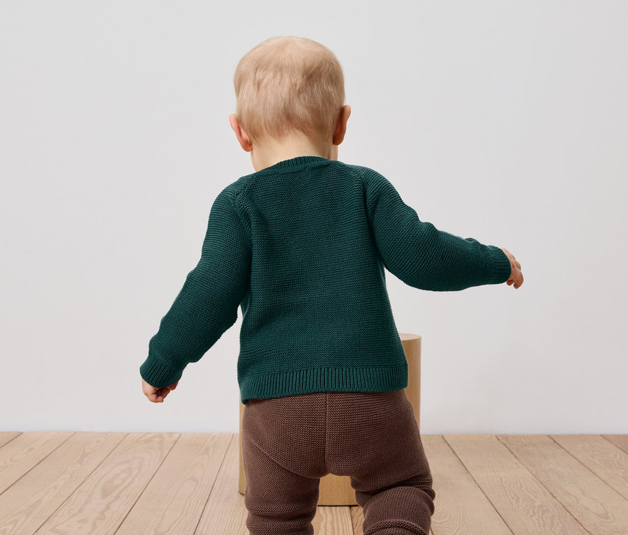 Un bébé portant un pull vert et des leggings marron, dos à la caméra.