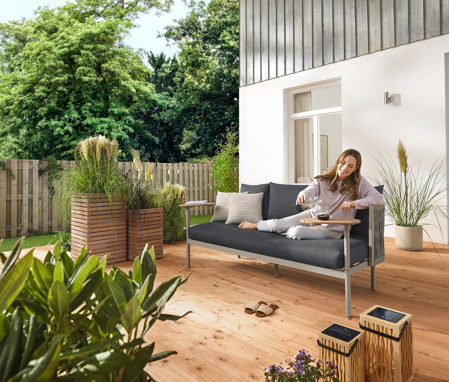 Une femme verse de l'eau dans un verre sur une terrasse de jardin. À côté d'elle se trouvent une petite et une grande lanterne solaire en bambou, 5 piquets à LED solaires et un canapé de jardin avec 2 tables tournantes et 2 colonnes à plantes « Tinus ».