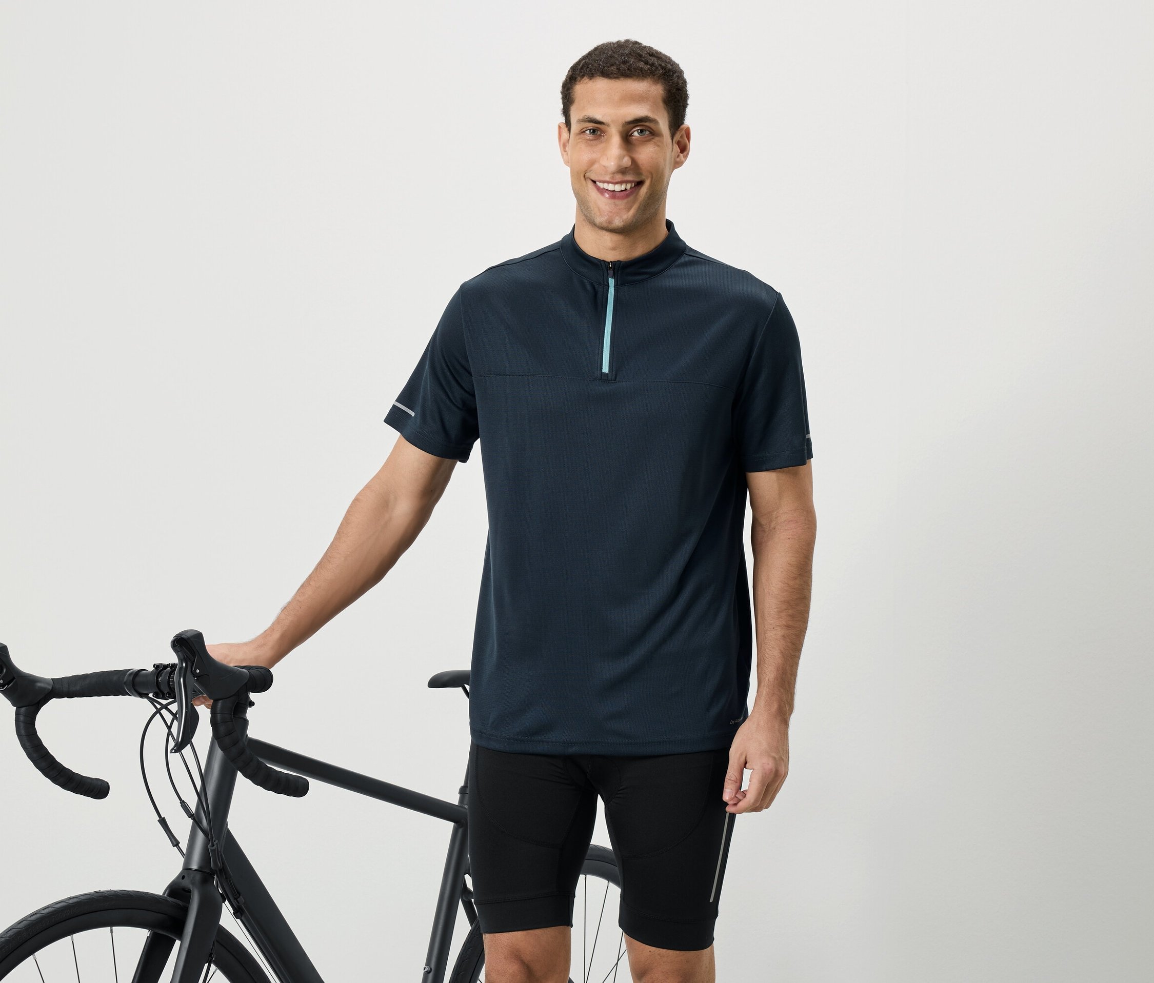 Un homme souriant portant une chemise à manches courtes bleu marine avec fermeture éclair se tient à côté d'un vélo.