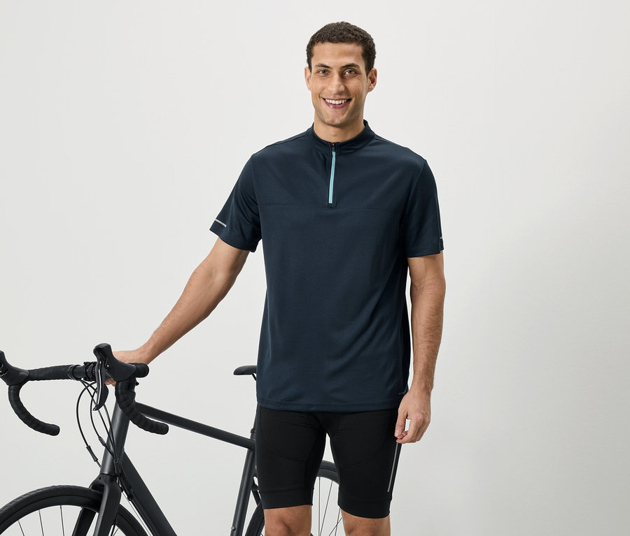 Un homme souriant portant une chemise à manches courtes bleu marine avec fermeture éclair se tient à côté d'un vélo.