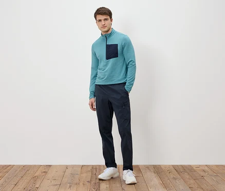 Un homme se tient devant un mur blanc, portant un T-shirt turquoise à manches longues avec une poche foncée, un pantalon bleu marine et des baskets blanches.