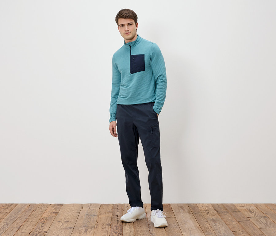 Un homme se tient devant un mur blanc, portant un T-shirt turquoise à manches longues avec une poche foncée, un pantalon bleu marine et des baskets blanches.