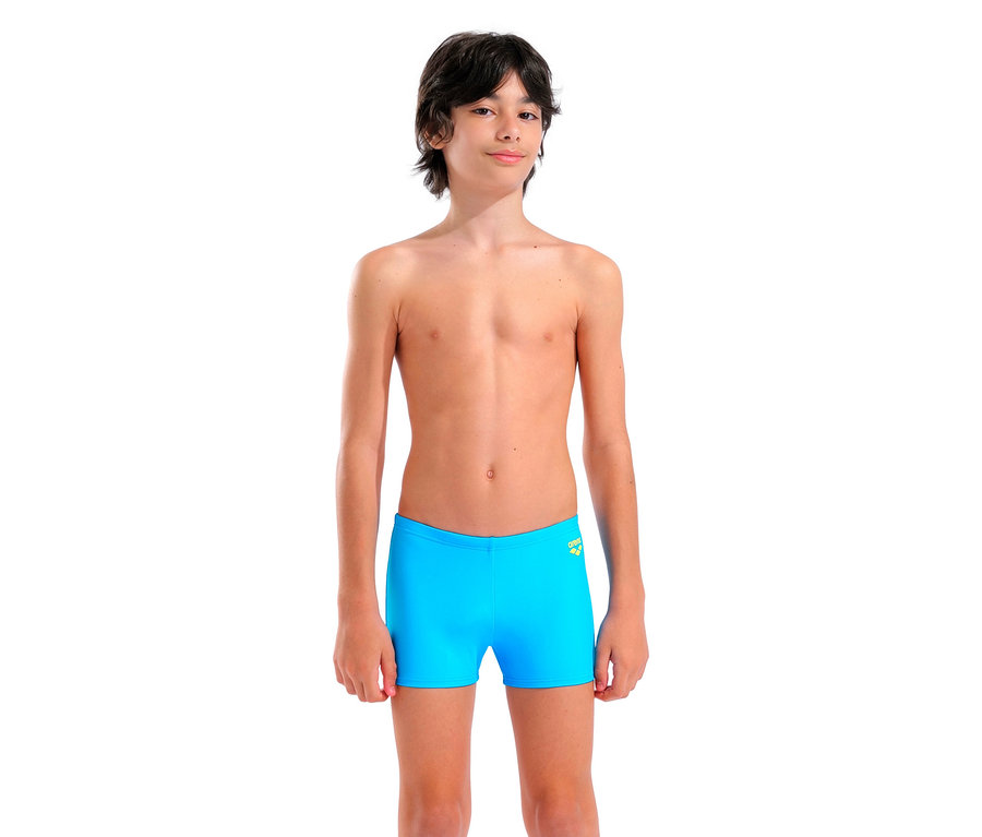 Un garçon se tient sur un fond blanc en maillot de bain bleu clair.