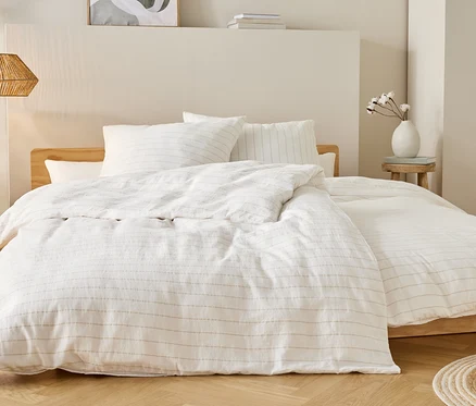 Lit avec des draps blancs à fines rayures, table de chevet avec vase et lampe suspendue au plafond.