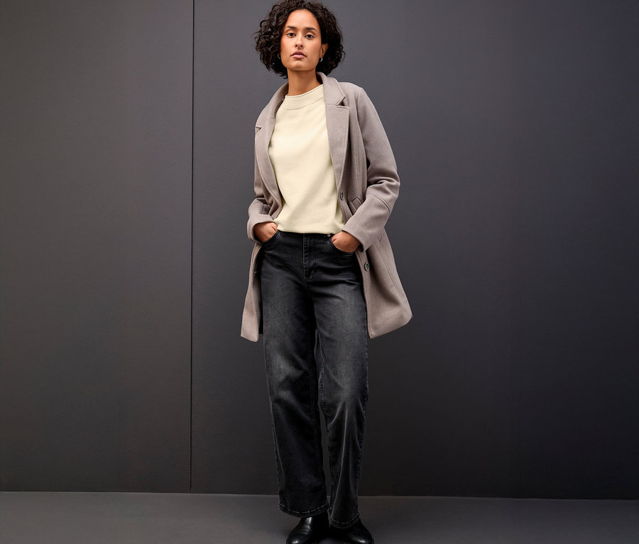 Une femme se tient debout, les mains dans les poches, portant un pull en tricot beige à bord roulé, une veste grise et un jean foncé.