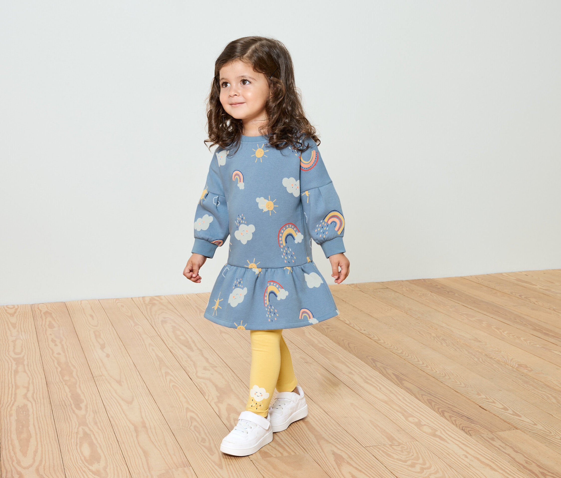 Une petite fille aux cheveux bouclés porte une robe bleue à motifs arc-en-ciel, des leggings jaunes et des chaussures blanches.