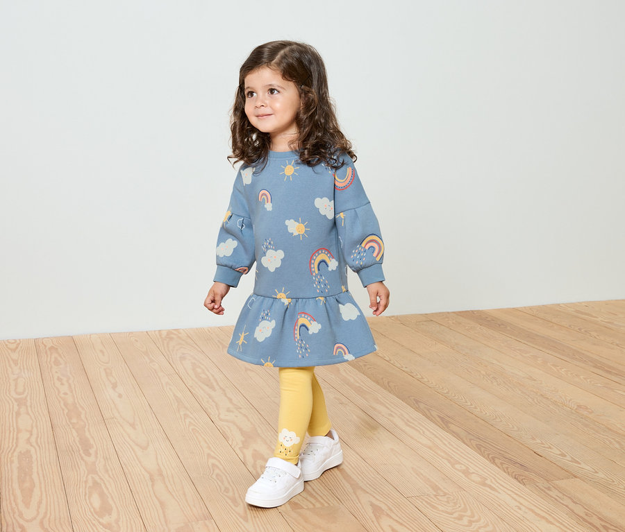Une petite fille aux cheveux bouclés porte une robe bleue à motifs arc-en-ciel, des leggings jaunes et des chaussures blanches.