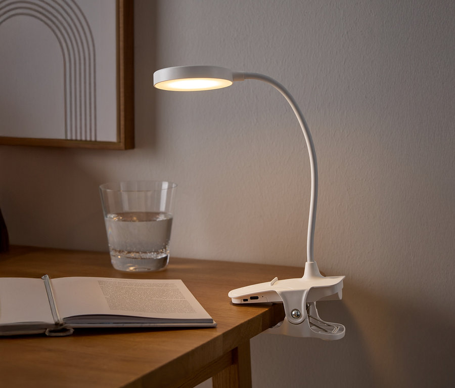 Une lampe à pince blanche illumine un livre ouvert sur une table en bois, à côté d'un verre d'eau et d'un tableau encadré.