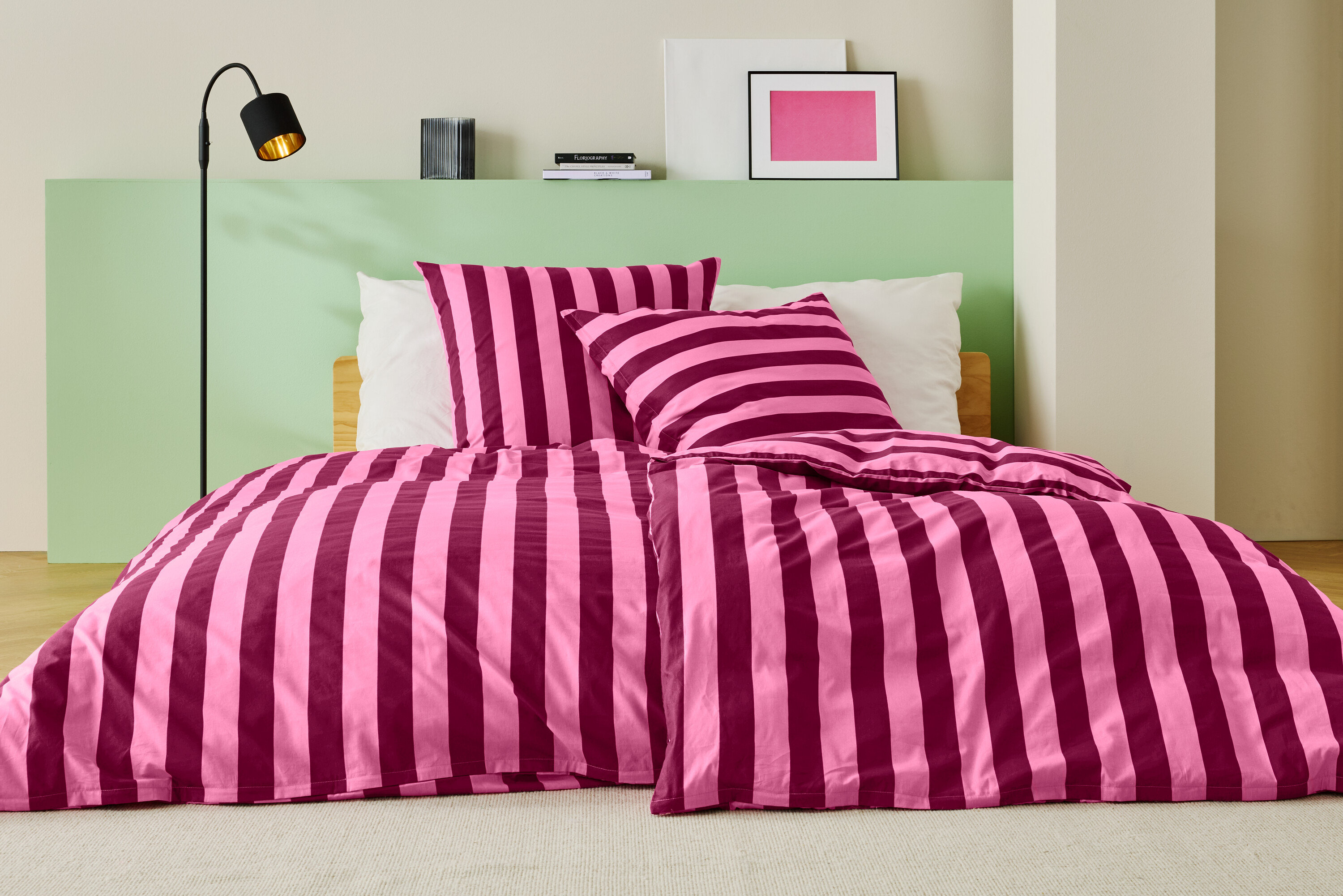 Lit avec des draps rayés rose et bordeaux, deux oreillers et un lampadaire.