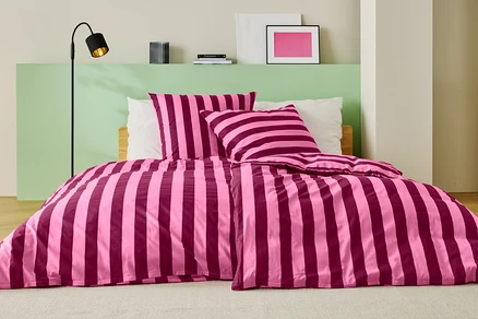 Lit avec des draps rayés rose et bordeaux, deux oreillers et un lampadaire.
