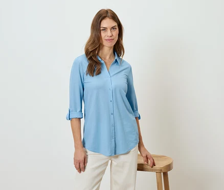 Une femme porte une chemise bleu clair et un pantalon beige, appuyée sur un tabouret.