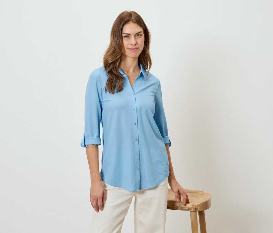 Une femme porte une chemise bleu clair et un pantalon beige, appuyée sur un tabouret.