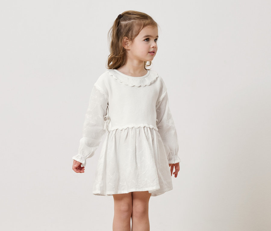 Une fille blonde porte une robe blanche à manches longues sur un fond neutre.