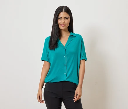 Une femme aux longs cheveux foncés porte un chemisier turquoise à manches courtes et un pantalon noir sur un fond clair.