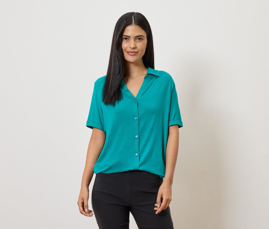 Une femme aux longs cheveux foncés porte un chemisier turquoise à manches courtes et un pantalon noir sur un fond clair.