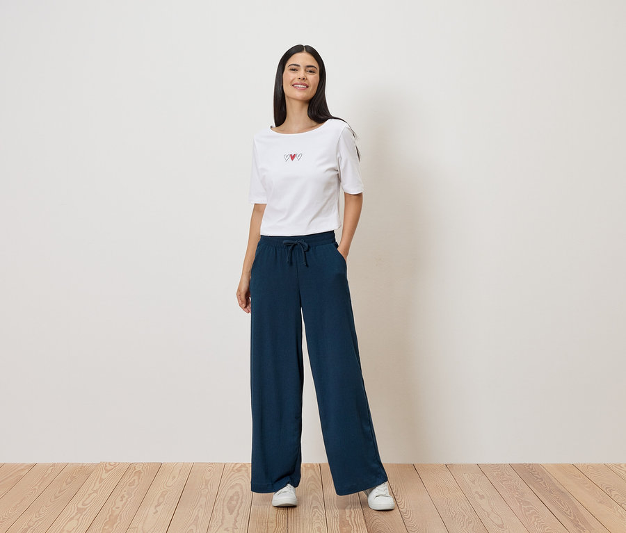 Une femme porte un t-shirt blanc avec un motif de cœur et un pantalon ample bleu marine à cordon de serrage. Elle porte des baskets blanches.