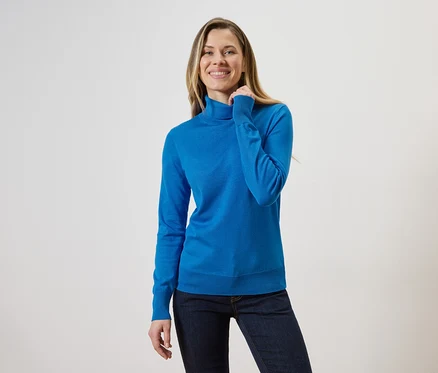 Une femme blonde portant un pull à col roulé bleu et un jean sur un fond clair.