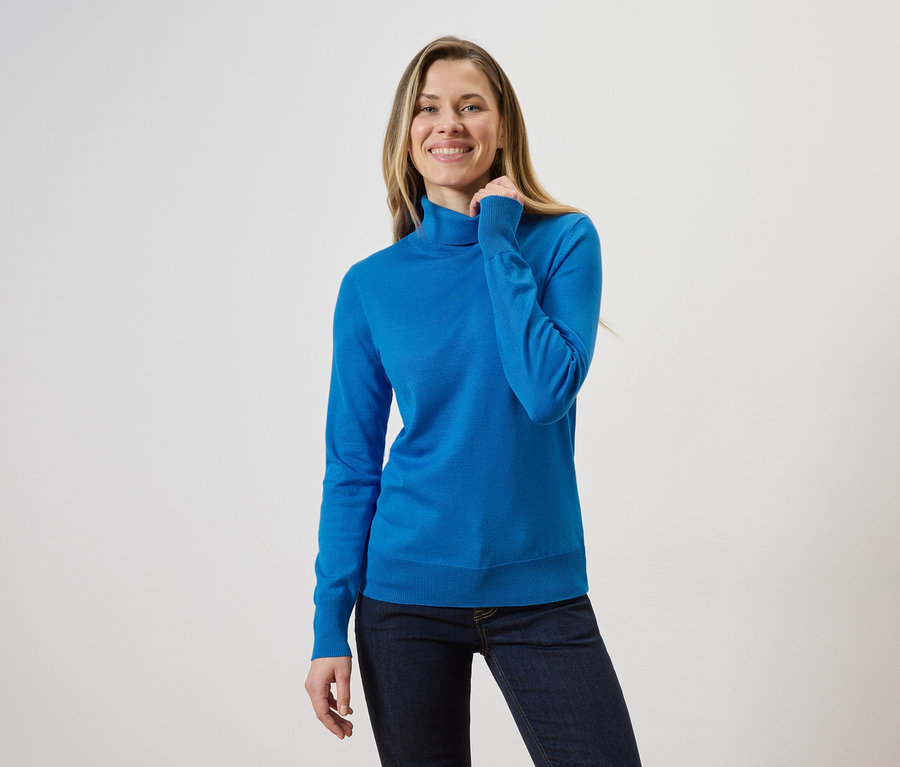Une femme blonde portant un pull à col roulé bleu et un jean sur un fond clair.