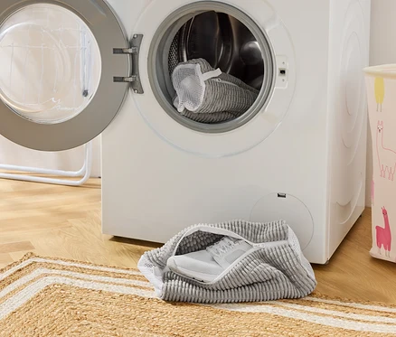 Un sac à chaussures gris avec une basket blanche est posé devant un lave-linge à porte ouverte. Un autre sac à chaussures se trouve dans le tambour.