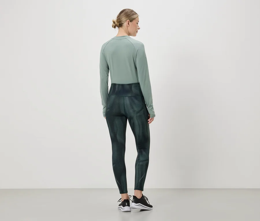 Une femme portant un chignon porte un haut à manches longues vert et un legging à motif abstrait, ainsi que des baskets noires.