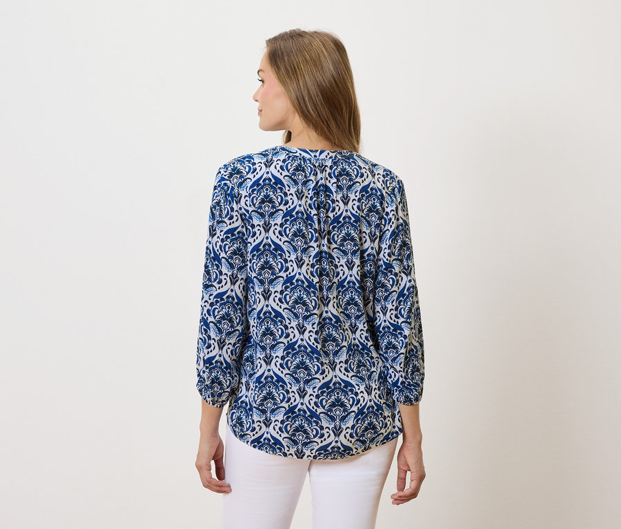 Une femme aux longs cheveux bruns porte un chemisier à motifs bleus et blancs et un pantalon blanc.