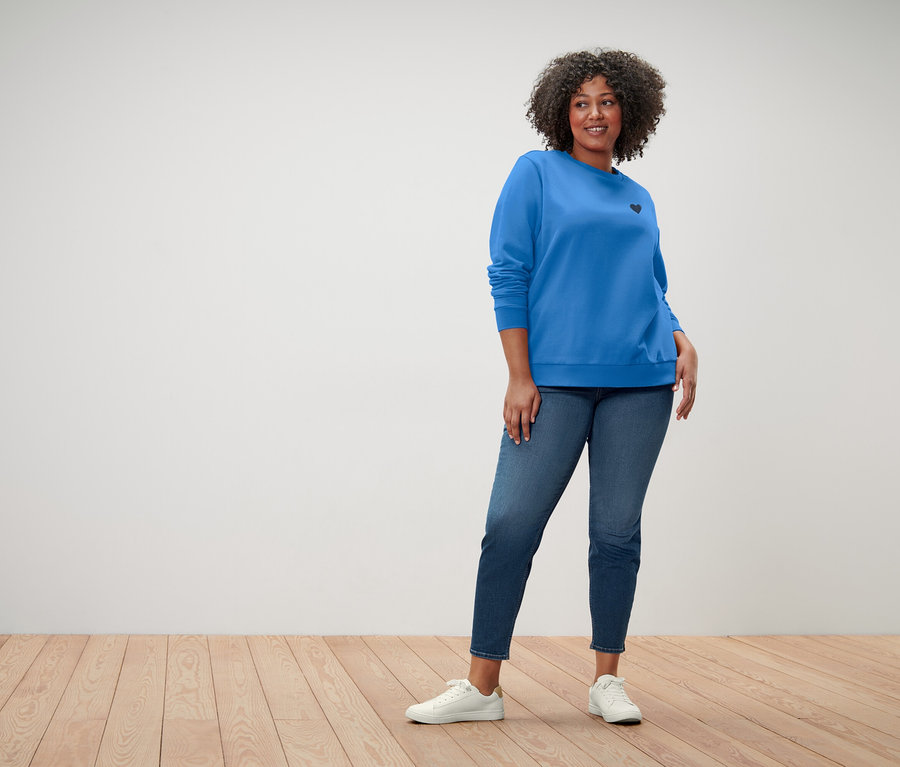 Une femme aux cheveux bouclés porte un pull bleu avec un petit cœur noir, un jean bleu et des baskets blanches.