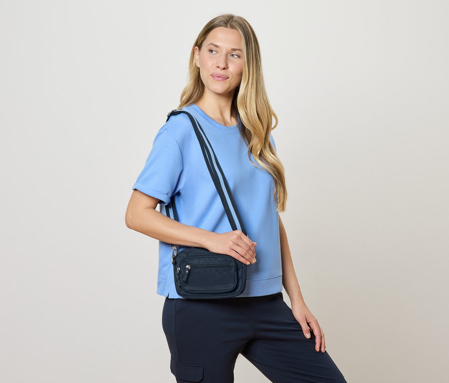 Une femme porte un sac bandoulière bleu marine avec des poches zippées, un haut bleu et un pantalon bleu marine.