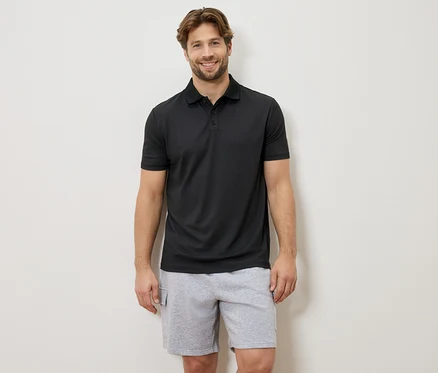 Un homme souriant se tient devant un mur, portant un polo noir et un short gris.