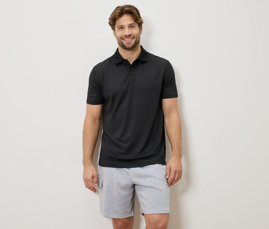 Un homme souriant se tient devant un mur, portant un polo noir et un short gris.