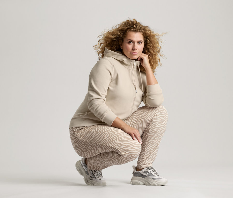 Femme aux cheveux bouclés, vêtue d'une veste beige, d'un pantalon à motif zébré et de baskets, accroupie avec la main sur le menton.