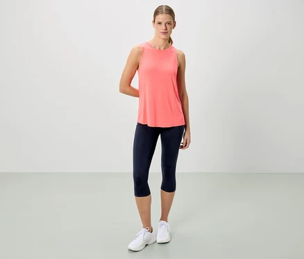 Une femme se tient devant un fond neutre en débardeur corail, legging capri bleu marine et baskets blanches.