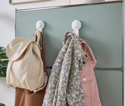 Un sac en tissu beige, un sac en daim marron, une chemise fleurie bleue et une veste en jean rose sont accrochés à des crochets à ventouse blancs sur un mur de placard vert menthe.