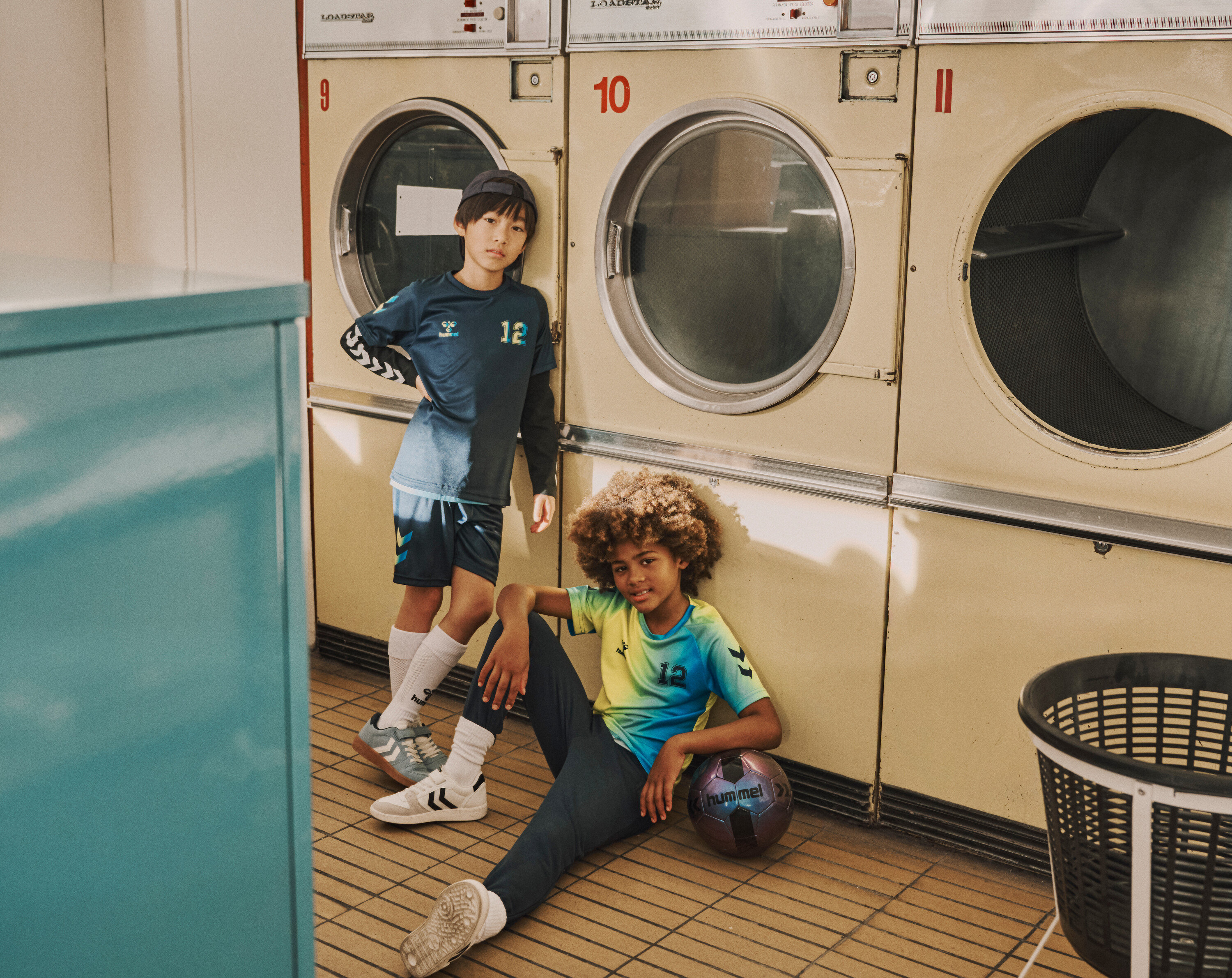 Deux garçons posent dans une laverie, l'un debout et l'autre assis avec un ballon de foot.