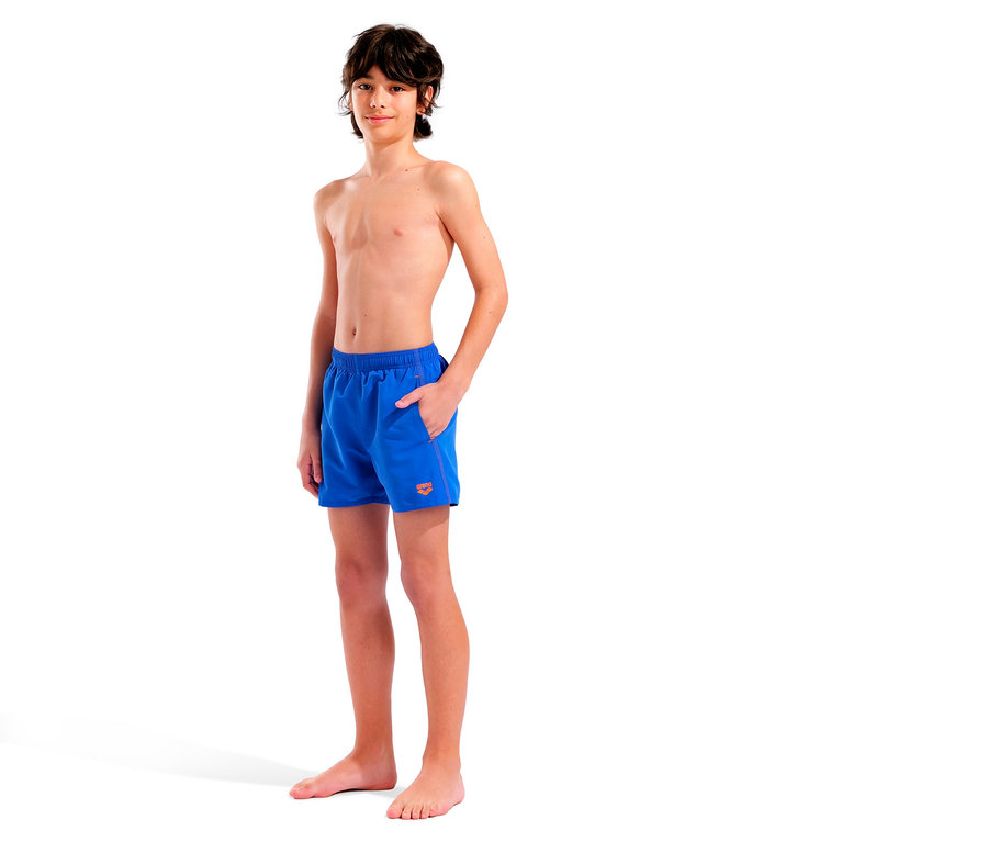 Un jeune garçon aux cheveux bruns, pieds nus, se tient sur un fond blanc en short de bain bleu avec un logo orange.