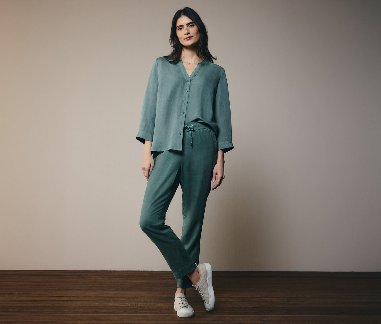 Une femme portant un chemisier boutonné vert et un pantalon, une jambe croisée sur l'autre, avec des chaussures crème.