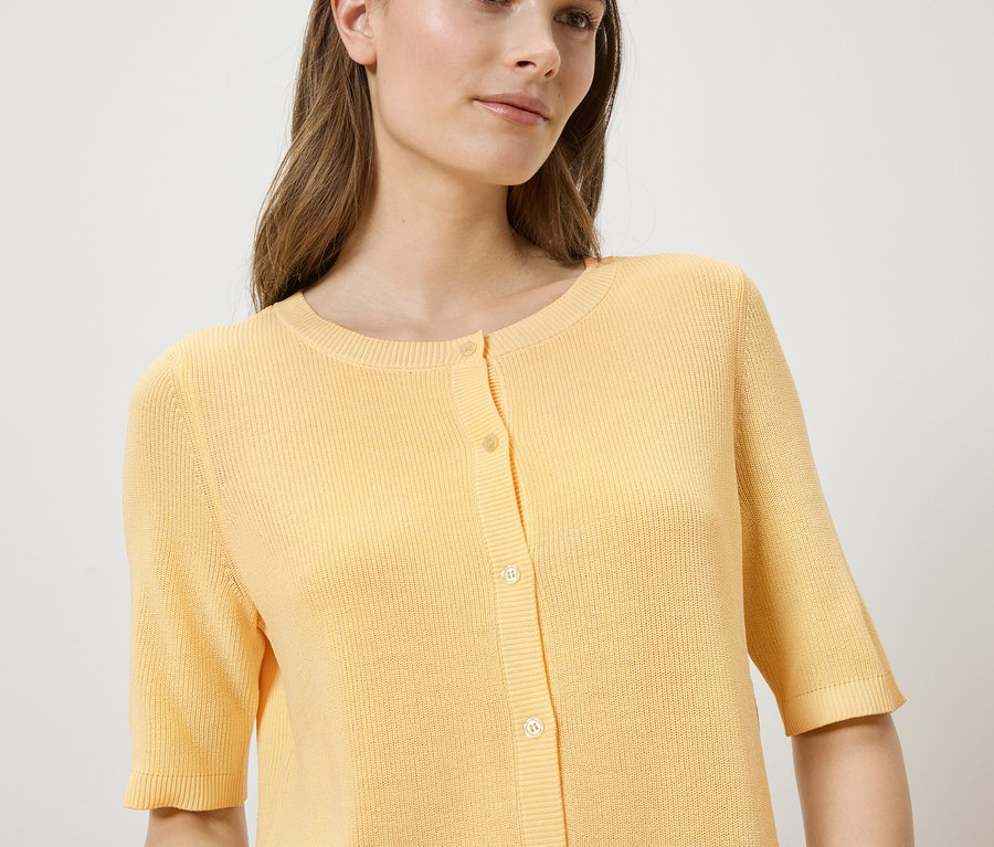 Une femme porte un cardigan jaune clair à manches courtes et à boutons sur un fond clair.