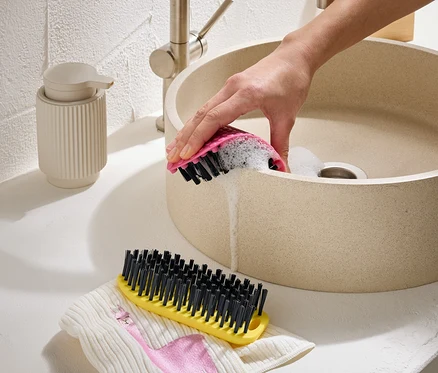 Une main nettoie un lavabo beige avec une brosse rose. Une brosse jaune et un tissu avec un flamant rose sont à côté.